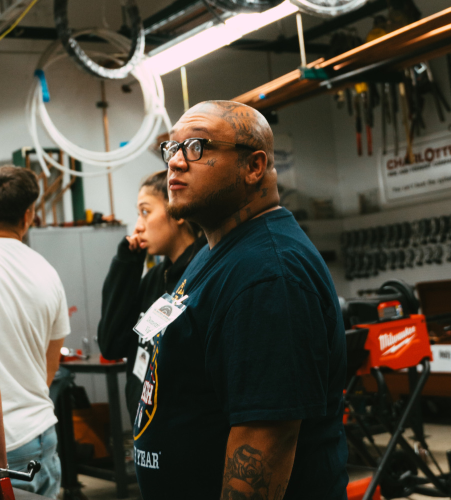 A group of people, including one with tattoos and glasses, stands in a workshop with various tools and equipment.